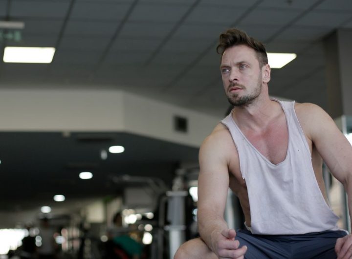 Muscular man with facial hair sitting and looking away in a gym, captured in selective focus.